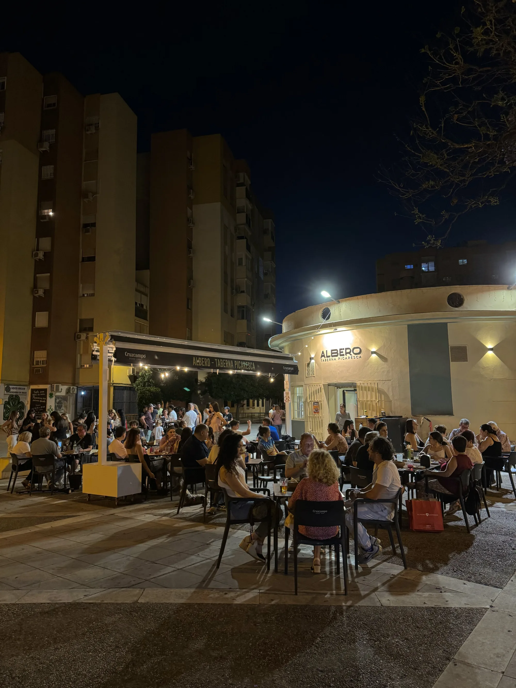 Terraza de Albero llena de gente por la noche, con el toldo de Cruzcampo iluminado